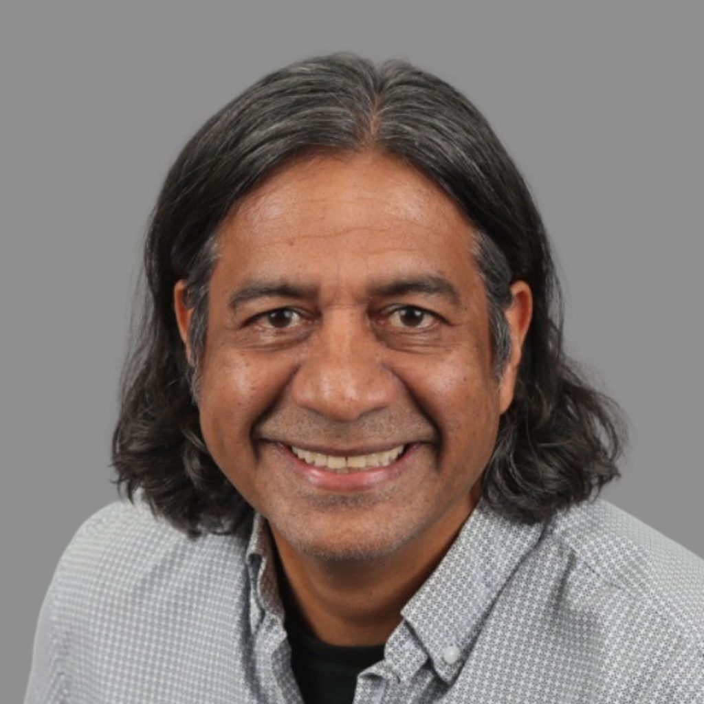 Professional headshot of Hugh Khan, featuring a warm smile and business casual attire against a neutral gray background.